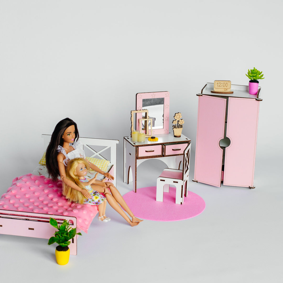 A miniature dollhouse bedroom set featuring a pink bed with a woman and child doll sitting on it, a pink vanity desk with mirror and accessories, a pink cabinet, and a pink rug, displayed on a light gray background.