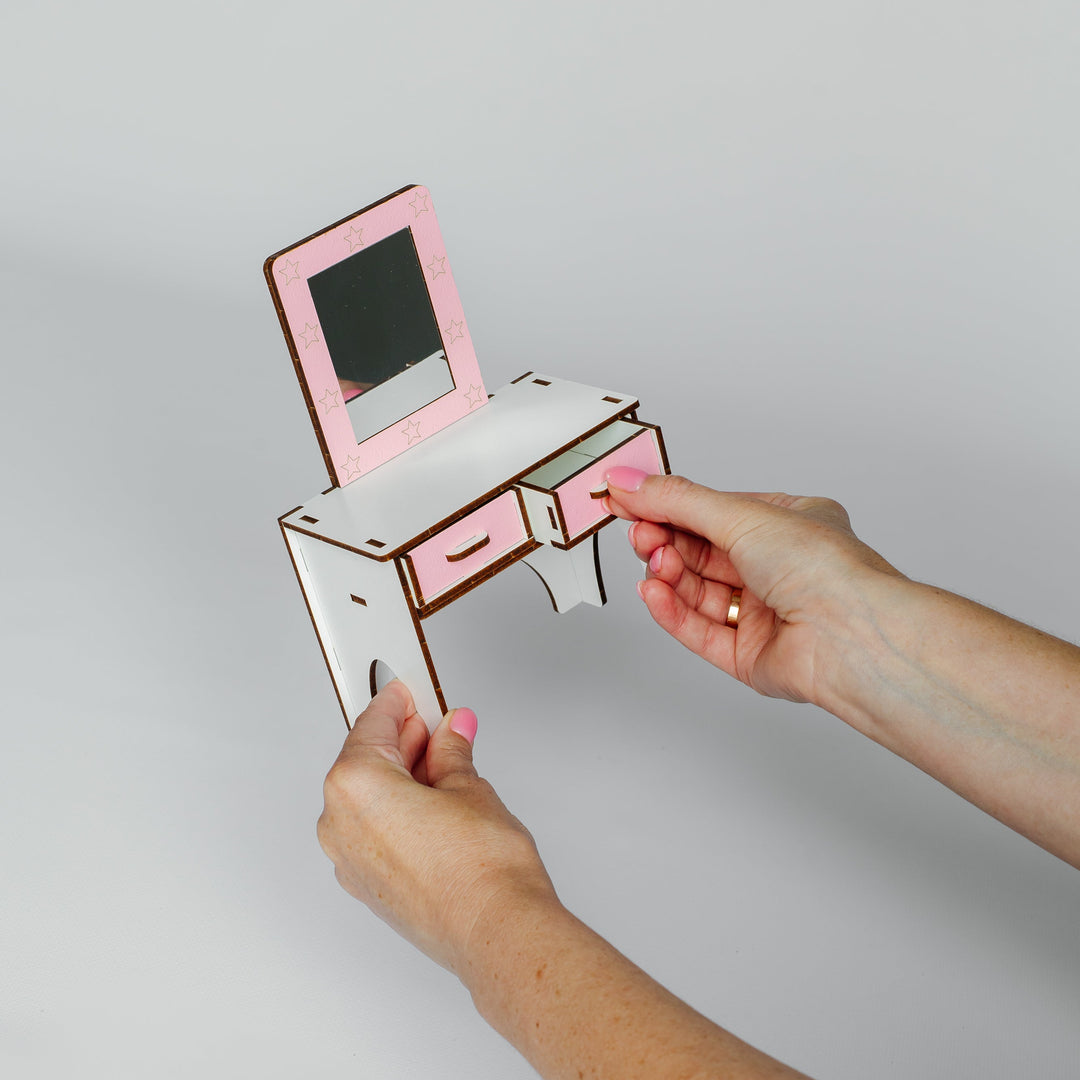 A person's hands holding a small pink and white wooden vanity desk with an attached mirror, shown against a plain light gray background.