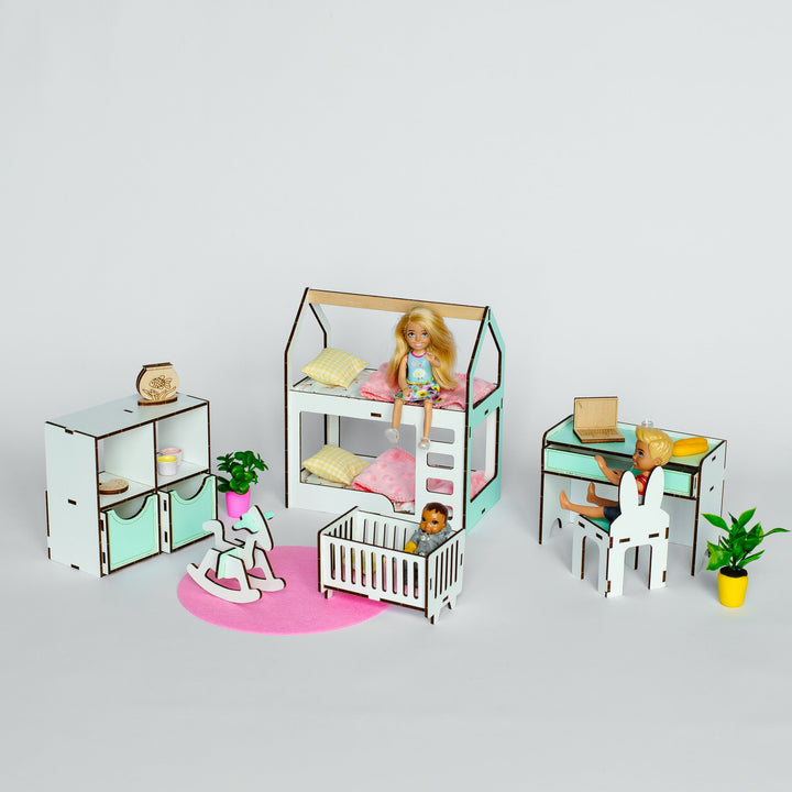 A dollhouse-style nursery furniture set displayed on a light gray studio background. The set includes a house-shaped bed with a doll inside, a crib, a small desk with a seated doll, a cabinet, a rocking horse, and a pink rug.