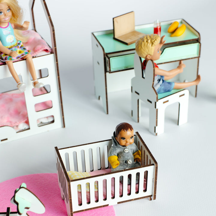 A miniature dollhouse nursery scene with small toy figures: one baby figure sitting in a crib, a seated child figure at a desk, and another figure on a bed. The scene includes a pink rocking horse on a pink rug against a light gray background.