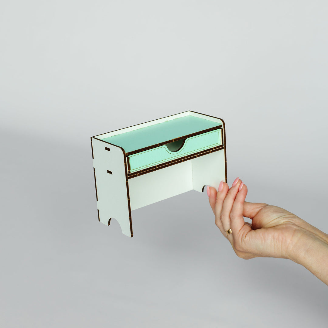 A small white and mint green wooden organizer drawer with a dark metal handle, held by a person's hand against a plain light gray background.