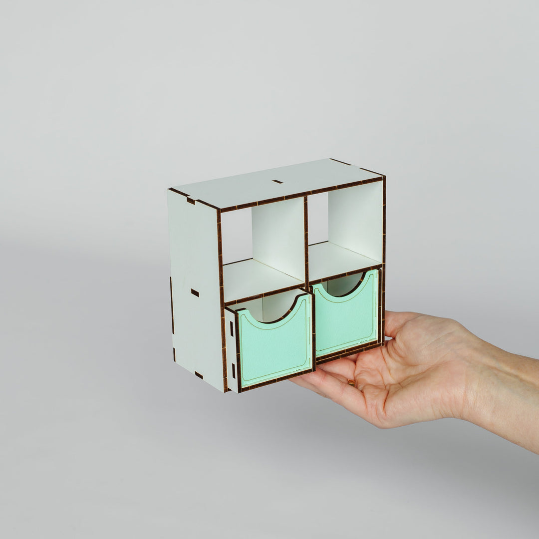 A hand holding a small laser-cut wooden organizer with two mint green drawer fronts and open upper compartments, set against a plain light gray background.