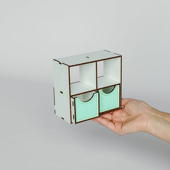 A hand holding a small laser-cut wooden organizer with two mint green drawer fronts and open upper compartments, set against a plain light gray background.