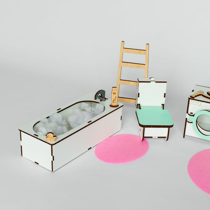 A miniature dollhouse bathroom set featuring a white bathtub with gray bubbly water, a light wood ladder, a mint green and white chair, and a matching vanity with a round mirror, arranged on a light gray background with pink circular mats.