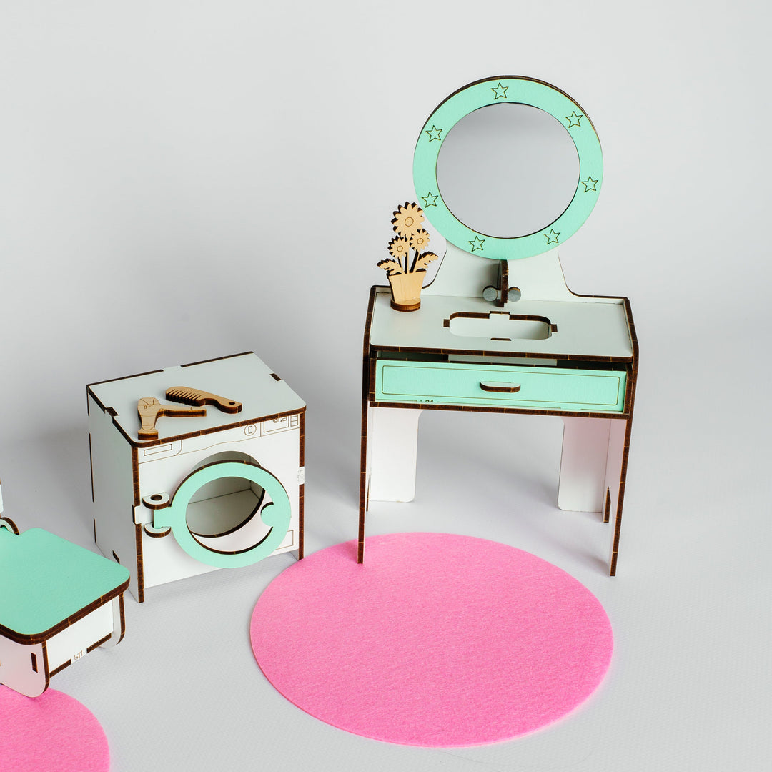 A miniature dollhouse-style room set with a vanity table, round mirror, washing machine, and small accessories, arranged on a light gray background with pink circular rugs.