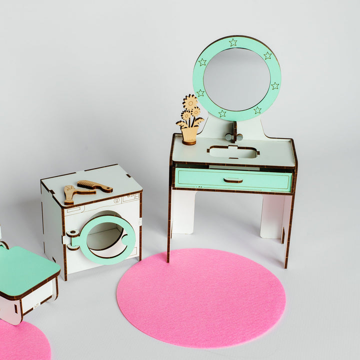 A miniature dollhouse-style room set with a vanity table, round mirror, washing machine, and small accessories, arranged on a light gray background with pink circular rugs.