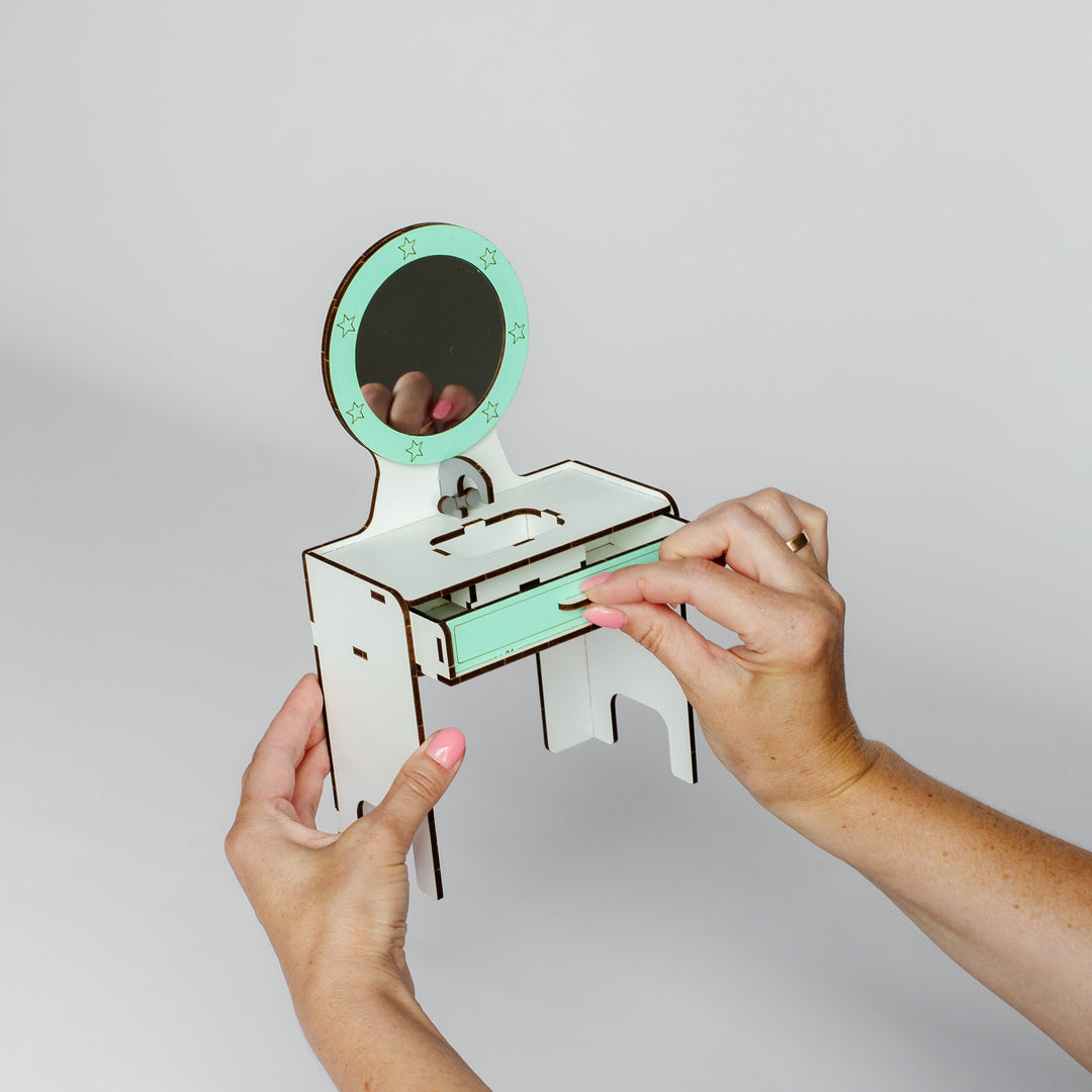 A person's hands holding a small wooden vanity desk with a mint green drawer and a round mirror, set against a plain light gray background.