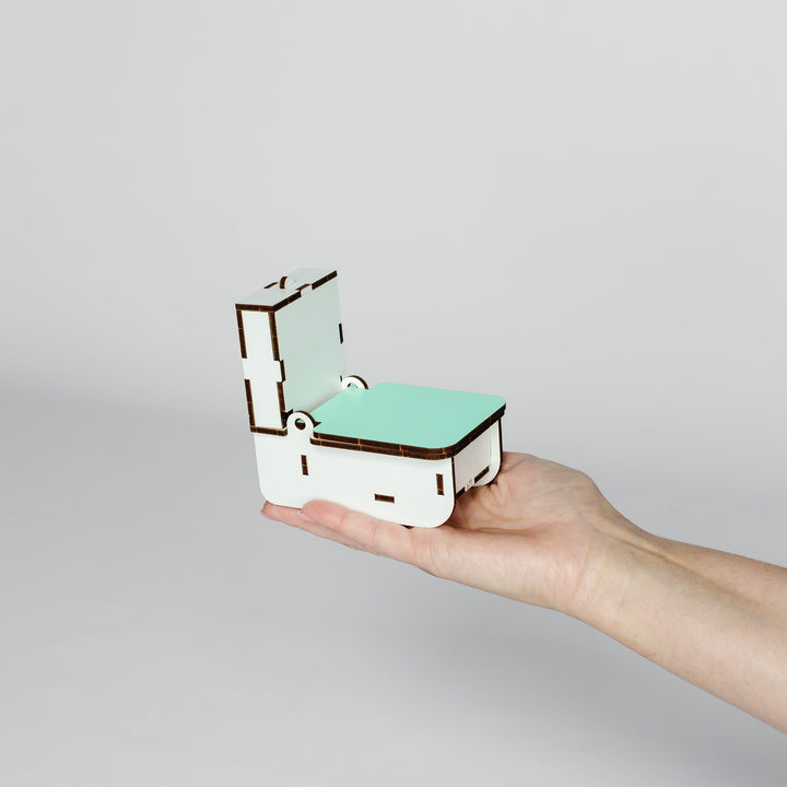 A miniature white toilet model with a mint green seat and dark brown laser-cut edges, held in the palm of a hand against a light gray background.