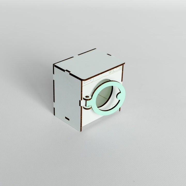 A miniature white laser-cut wooden toy washing machine with a mint green circular door, shown on a neutral light gray background.