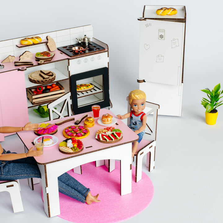 A miniature cardboard kitchen playset with a dining table, stove, refrigerator, and accessories, shown with small doll figures and a child-like figure in a bright studio setup.
