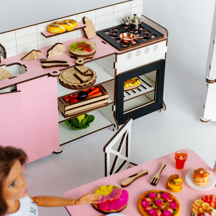 A dollhouse-style pink kitchen set with miniature food items, a stove, oven, counter, and dining table, shown with a doll in the foreground against a clean white background.