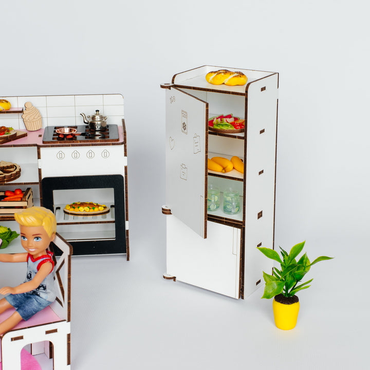 A miniature dollhouse kitchen scene with a white and brown toy refrigerator stocked with food, a small stove, and a doll seated at a table, photographed on a clean light gray studio background.