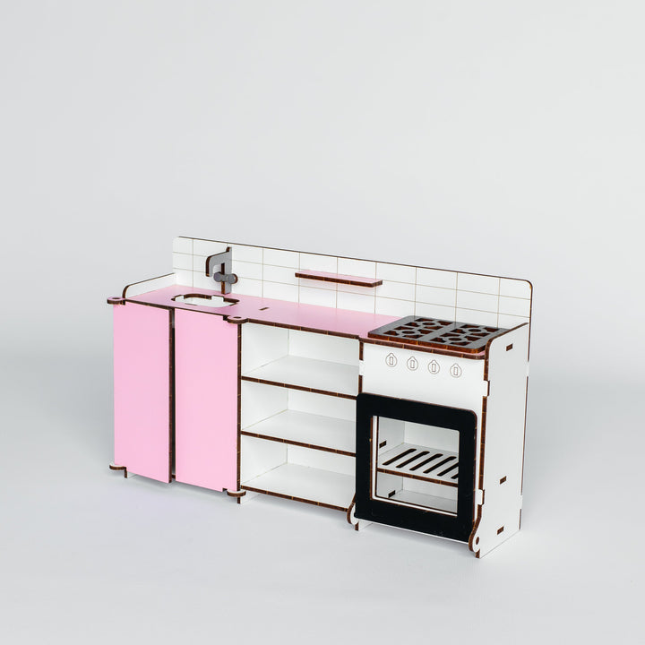 A miniature toy kitchen set with pink cabinets, a white tiled backsplash, a black stovetop, and a glass-front oven, photographed on a seamless light gray background.
