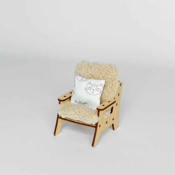 A miniature wooden armchair with a fluffy cream sheepskin-style seat and back cushion, plus a small white decorative pillow, photographed on a seamless light gray studio background.