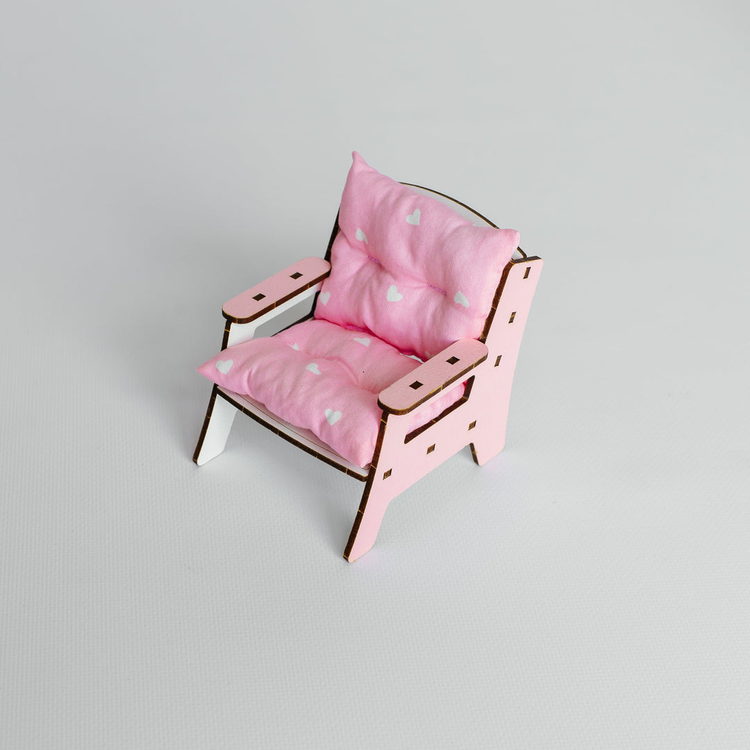 A miniature pink doll chair with a matching pink cushion, featuring a light wood frame and small white feet, set against a plain light gray background.