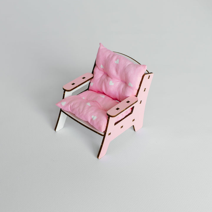 A miniature pink doll chair with a matching pink cushion, featuring a light wood frame and small white feet, set against a plain light gray background.