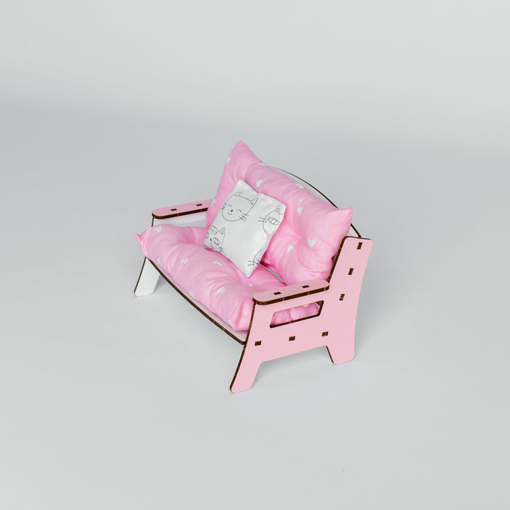 A miniature pink dollhouse sofa with patterned pink cushions and a small white accent pillow, shown on a light gray studio background.
