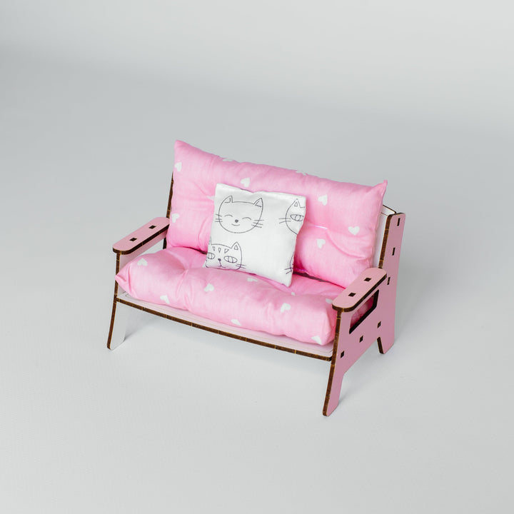 A miniature pink loveseat with a wooden frame, featuring pink cushions with white polka dots and a small white accent pillow with a black cat illustration, set against a plain light gray background.