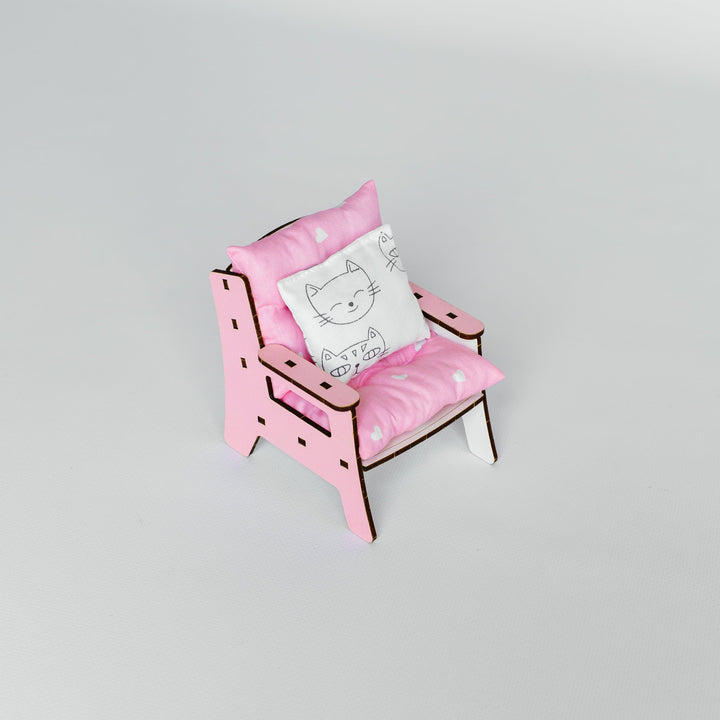 A miniature pink wooden chair with matching pink cushions and a small white decorative pillow featuring a black cat illustration, set against a plain light gray background.