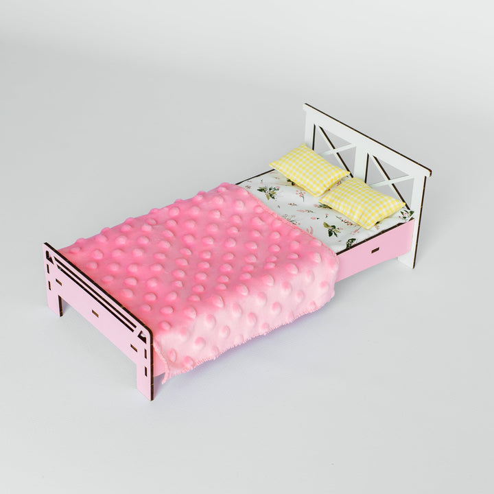 A miniature pink wooden dollhouse bed with a textured pink quilt, yellow pillows, and a white headboard, shown on a light gray background.