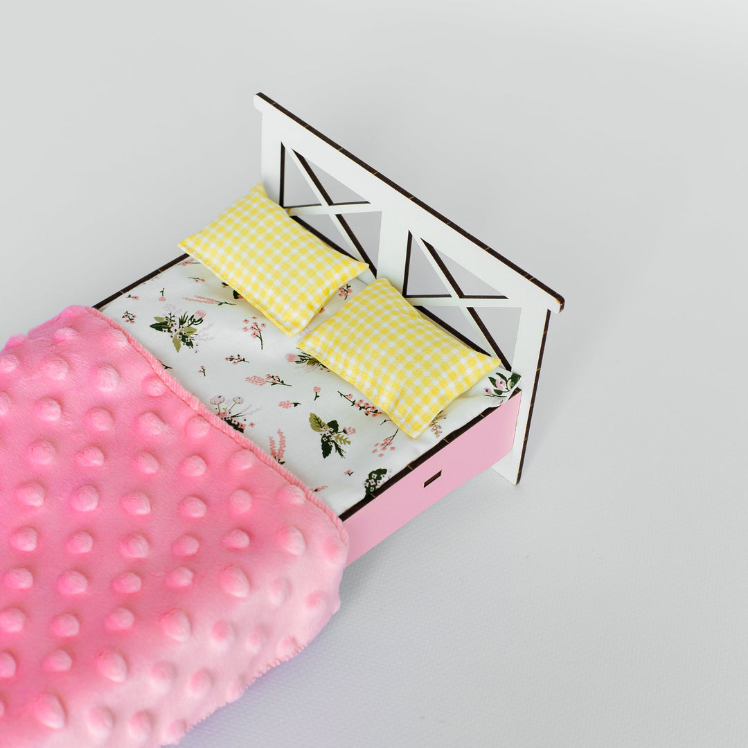 A miniature dollhouse bed with a pink frame, white headboard, yellow gingham pillows, and a pink textured blanket on a light gray background.