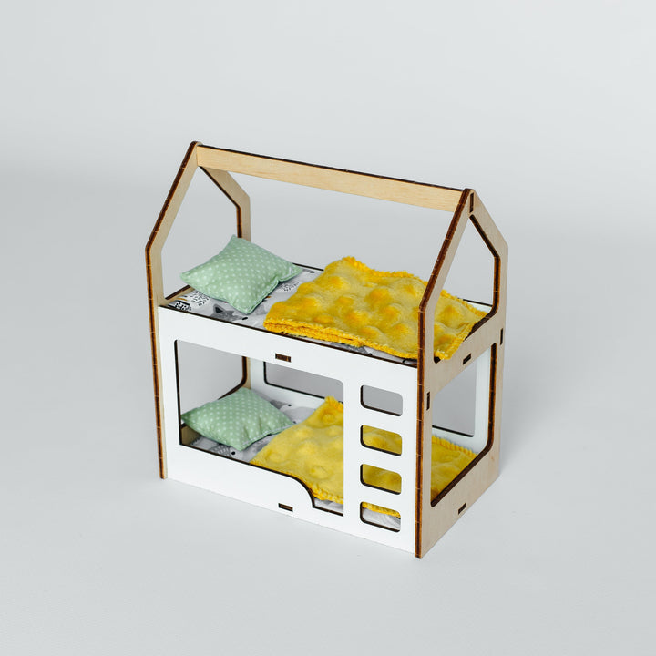 A miniature laser-cut wooden house-shaped bed frame with two levels, styled with yellow blankets and mint green pillows on a light gray background.