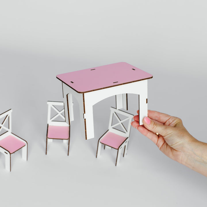 A miniature wooden dollhouse dining set with a pink table and white chairs with pink seats, shown with a hand holding one of the chair legs against a light gray background.