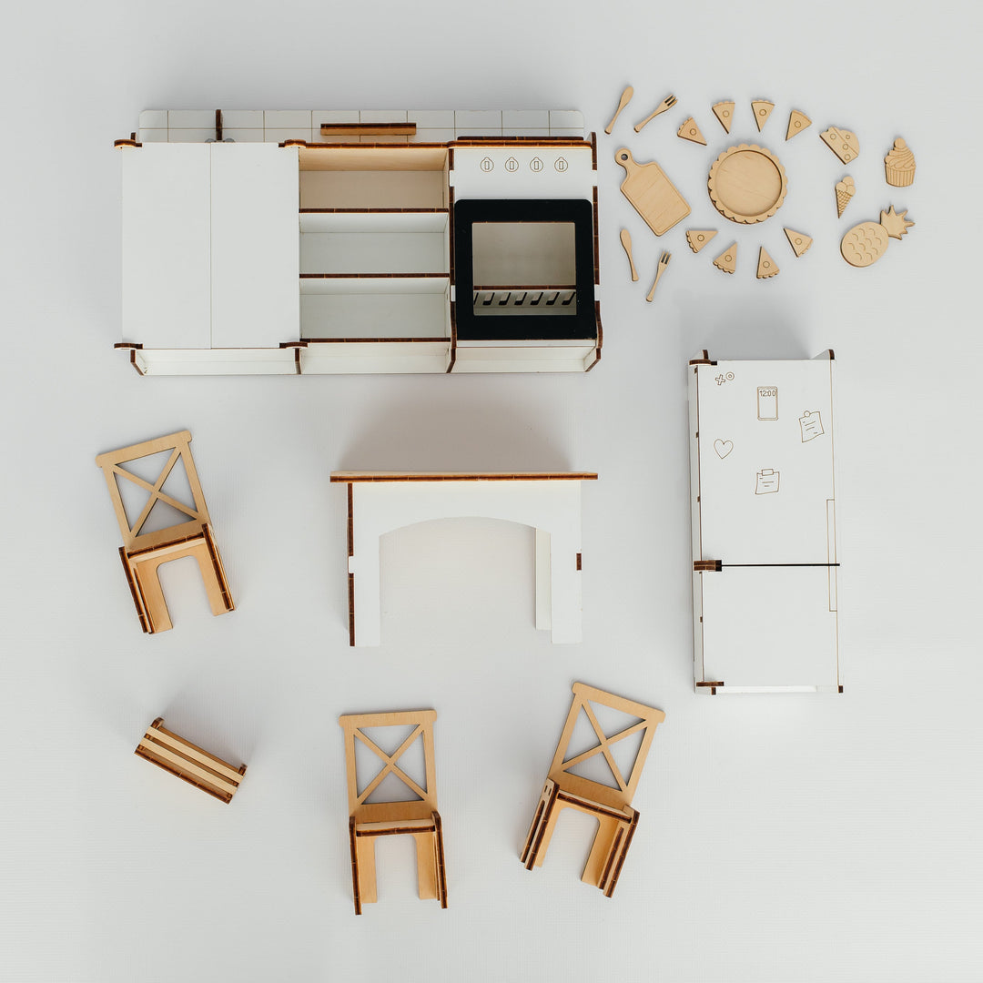 A top-down view of a miniature wooden kitchen furniture set, including a white and wood cabinet unit, stove, refrigerator, table, chairs, and decorative wooden pieces, arranged on a clean white background.