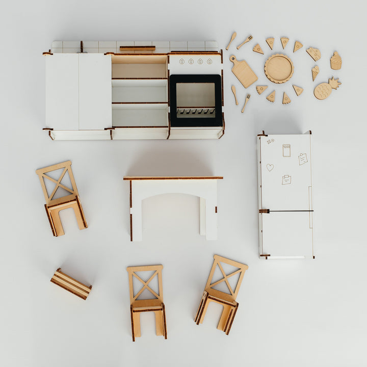 A top-down view of a miniature wooden kitchen furniture set, including a white and wood cabinet unit, stove, refrigerator, table, chairs, and decorative wooden pieces, arranged on a clean white background.