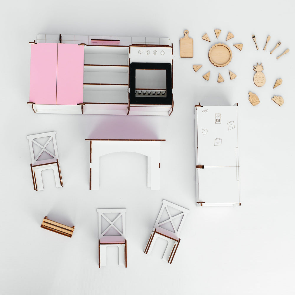 A miniature dollhouse kitchen set arranged on a white background, featuring a pink and white kitchen unit with a black stove, a white refrigerator, several small chairs, and wooden serving pieces and utensils.