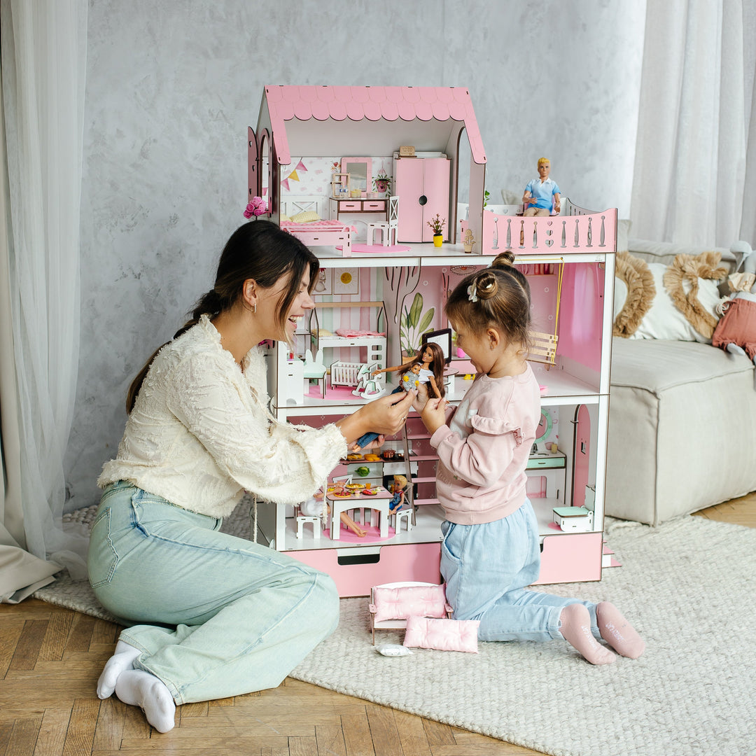 Woman and child playing with a pink dollhouse in a room with a gray wall and wooden floor.