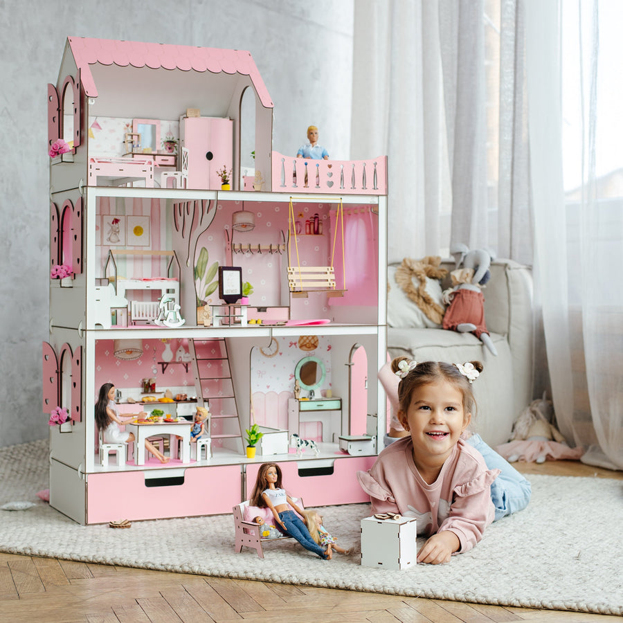 A young girl smiling on a white rug in a bright room, playing with a large pink and white dollhouse filled with miniature furniture and dolls.