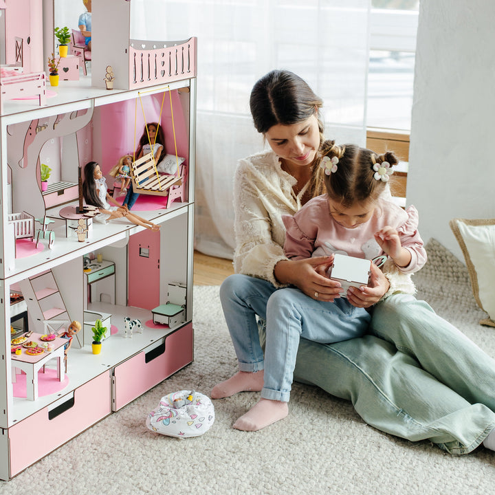 Woman and child playing with a pink dollhouse in a bright room.