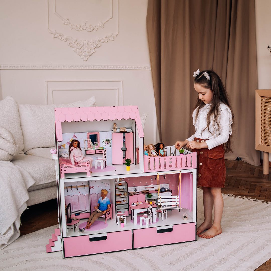 Young girl playing with a pink dollhouse in a room with wooden flooring and a cabinet.