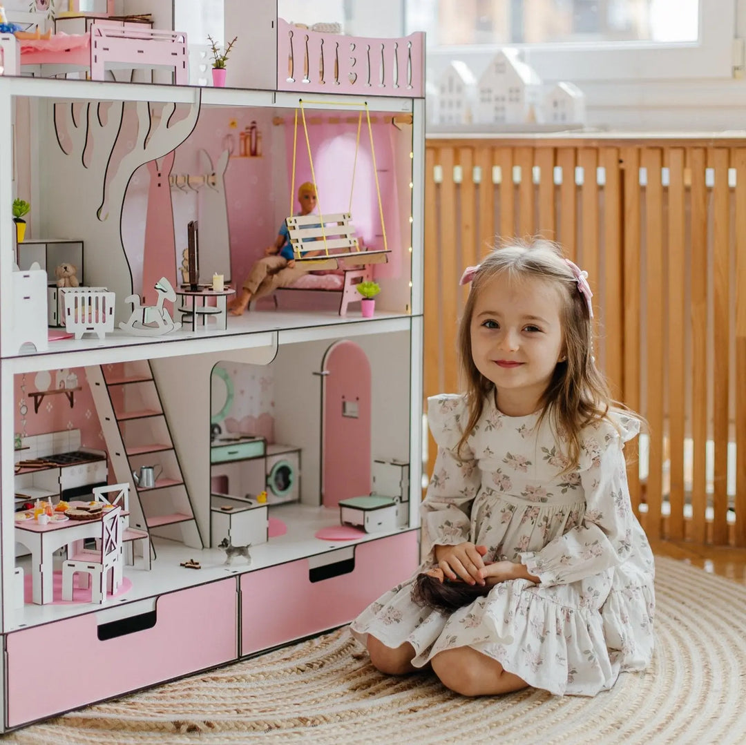 Young girl sitting next to a pink dollhouse with a doll inside.