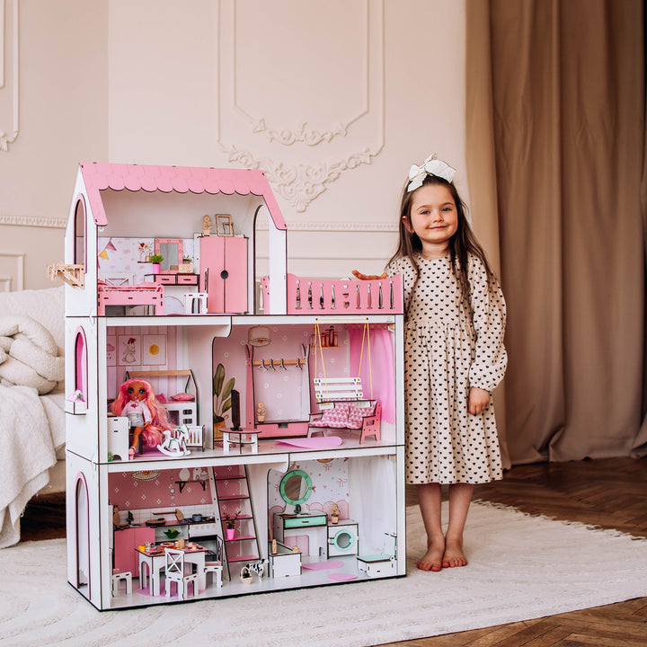 Young girl standing next to a large pink dollhouse in a room with beige curtains and wooden floor.