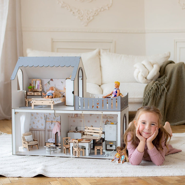 A smiling girl lying on the floor next to a two-story wooden dollhouse with furniture and dolls inside. The dollhouse is painted in white and natural wood colors, featuring a balcony.