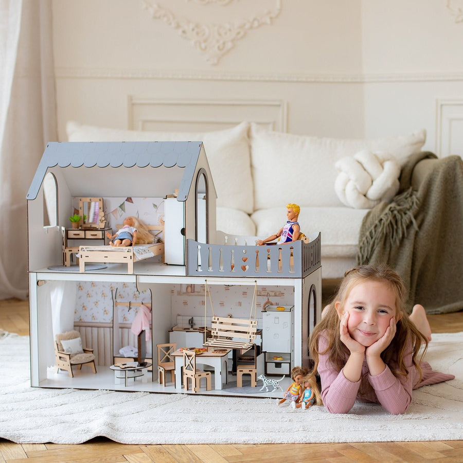 A smiling girl lying on the floor next to a two-story wooden dollhouse with furniture and dolls inside. The dollhouse is painted in white and natural wood colors, featuring a balcony.
