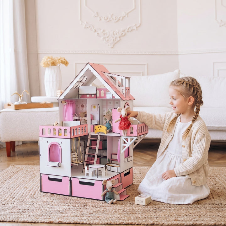 A young girl with braided hair sits on a woven rug in a bright living room, interacting with a large pink and white wooden dollhouse. The dollhouse features a detailed interior with miniature furniture and stairs.