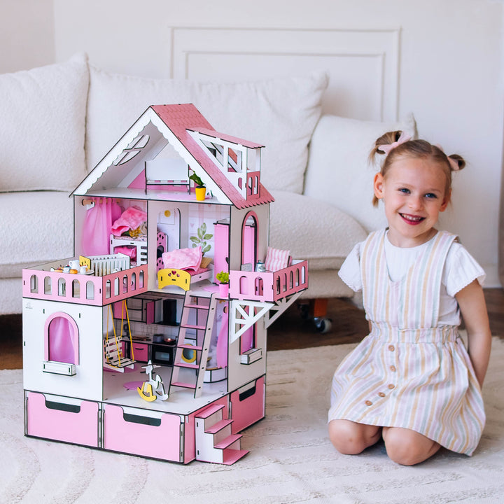 A young girl smiling and sitting next to a wooden dollhouse with a pink and white color scheme. The dollhouse is multi-storied with various rooms and a swing outside.